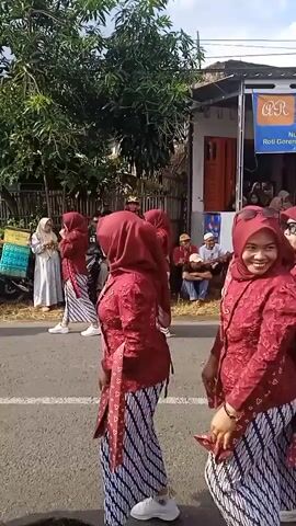 The video shows a group of women performing a dance routine in the street.  They are wearing matching outfits of red tops with ornate patterns, striped batik-style skirts, and red hijabs. They move in unison with controlled steps and hand gestures.  The dance occurs on a paved road in what appears to be a residential neighborhood.  A crowd of spectators sits along the side of the road, watching the performance. Behind the crowd is a building with a banner that has text in Indonesian that translates to "Selling Nugget Karage, Sausage, Soslis, Fried Bread," and mentions an address.  There are trees and parked motorcycles also visible. The lighting is bright indicating an outdoor daytime setting.
