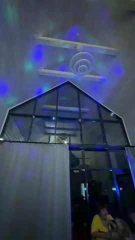 A low-angle, wide shot captures a room with a high, sloped ceiling made of wood planks. A modern, multi-tiered white light fixture hangs from the center of the ceiling. Below the fixture, a large, triangular window spans the upper portion of the room, revealing the roof structure behind it. Colored lights, likely from a disco ball or similar effect, are projected onto the ceiling and window, creating vibrant green, blue, and purple spots. A sheer white curtain hangs from the left side of the frame, partially obscuring the view into the upper part of the room. In the foreground, partially visible on the right, are several people sitting at a table. One person wearing a yellow top is facing the camera and smiling. The overall atmosphere suggests a social gathering or party.