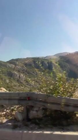A car is traveling on a road, likely in a rural or mountainous area. The camera is positioned to capture the view through the car's window, showing a landscape of rolling hills and mountains covered in green vegetation. There are some small buildings or houses visible in the valley between the hills. A metal guardrail is in the foreground, and the movement of the car is evident in the motion blur of the surrounding foliage. The sky is clear and blue, with some sun glare reflecting on the window.