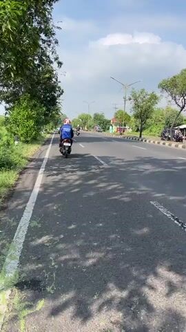 The video captures a daytime scene on a paved road, likely in a rural or suburban area given the presence of trees and open sky. The road has two lanes and a white dotted line dividing them. Shadows of trees are cast across the road. On the left side of the road, a person on a white and blue motorcycle is riding away from the camera, wearing a blue jacket and a white helmet. Further down the road, another motorcycle is visible. On the right side of the road, a black SUV is parked, and another black vehicle is partially visible behind it. The environment appears sunny with some clouds in the sky.