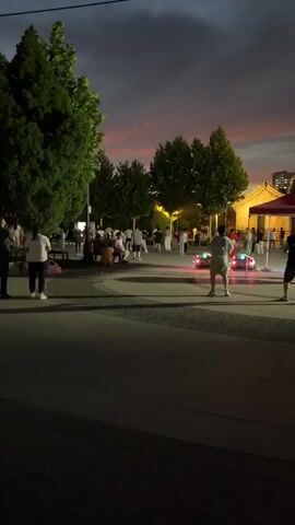 A group of people are gathered outdoors at dusk. Most are standing and some are seated on a bench. Several individuals are dancing in the middle of the paved area. A person in the foreground on the right is facing away from the camera and appears to be holding a device that emits pink and blue lights. A small, red object with lights is on the ground in the middle ground to the right of the dancing people. Trees and some artificial lighting are visible in the background. The sky is a deep purple and pink, indicating sunset or twilight.