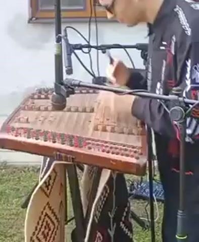 A person wearing glasses and a black shirt with white embroidery is playing a dulcimer. The dulcimer is on a stand. Microphones are positioned above and to the side of the instrument, suggesting it is being amplified or recorded. The person is using small hammers to strike the strings. The background appears to be outdoors, with a building and grass visible. There are also some patterned cloths hanging from the stand. The scene is captured in a close-up shot focused on the musician and the instrument.