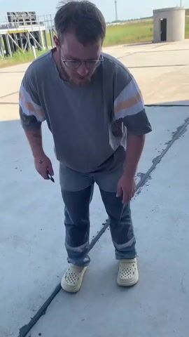 A man with short brown hair, glasses, and a beard stands in an outdoor area on a paved surface. He is wearing a gray t-shirt with horizontal stripes on the sleeves and dark pants with lighter patches. He is also wearing light-colored Croc-like shoes. He is holding a black object in his right hand. In the background, there is a metal structure with stairs, a small circular building, and a tall tower in the distance. The sky is blue with some clouds. The man appears to be looking off to his right and speaking.
