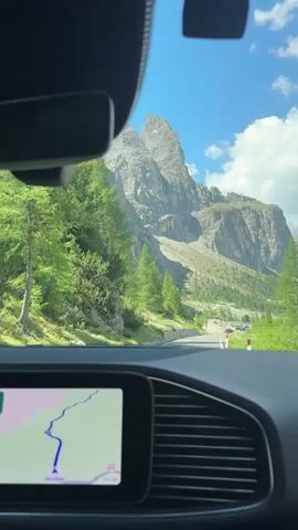 The video is shot from inside a car, looking through the windshield at a mountain landscape. The dashboard is visible in the foreground, with a navigation screen showing a route. The road ahead winds through a valley surrounded by steep, rocky mountains and green trees. Several cars are visible on the road in the distance. The sky is blue with scattered white clouds. The overall impression is of a scenic drive through a mountainous region on a sunny day.