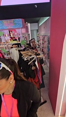 The video shows the interior of a bright pink makeup store. Customers shop for clothes and makeup at several display tables. A woman in pink stands near a round table with a mirror and makeup, while another woman examines clothes on a rack. Several customers look at makeup products in a pink display case against the wall. Other makeup products are neatly arranged on tables, and a wall reads "Sombre aqui + Sombre alla." The ceiling is decorated with a curved, white panel and a black panel with the word "LOVE" in graffiti style. The overall atmosphere is bright and cheerful.