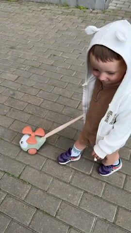 A young child, likely a toddler, is filmed walking on a paved sidewalk. The child wears a white hooded sweatshirt with bear ears on the hood, a brown shirt, brown shorts, and purple sneakers. The child is holding the handle of a toy scooter with a pink and green propeller. The camera appears to be held by an adult, and the perspective shifts between the child's face and an overhead view of the child's feet and the scooter. The child's gaze is directed downwards, suggesting concentration or curiosity. The setting appears to be outdoors, with the gray brick paving stones and dappled light indicating it could be daytime. The footage is in Russian, with the child or narrator asking "Where to go?" and then responding "There." The overall tone is lighthearted and focuses on the child's exploration and interaction with the toy.