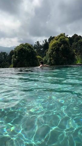The video shows a person swimming in an infinity pool. The person is seen from the back, with their head and shoulders above the water. They are wearing dark swimming trunks. The pool is surrounded by lush green trees and mountains in the background. The sky is cloudy, and the sun reflects on the water, creating a shimmering effect. The water in the pool is clear and blue-green. The person exits the pool by pushing up and out onto the edge.