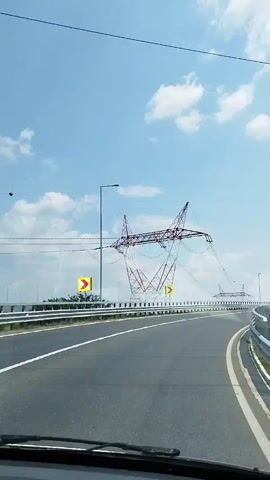 The video shows a view from inside a vehicle driving on a multi-lane highway under a bright blue sky with scattered white clouds. The highway has clear lane markings. On the left side of the highway, there are guardrails and a large electricity pylon. A yellow curved arrow sign is visible on the left. Further down the road, more electricity pylons can be seen. The camera is pointed forward, showing the road ahead and the sky above. The scene appears to be set in a rural or suburban area, judging by the open landscape visible beyond the highway.