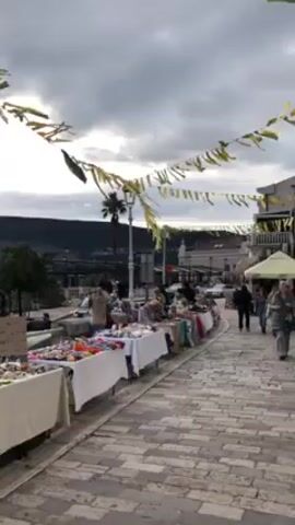 The camera pans across an outdoor marketplace on a cloudy day, with rows of tables displaying handmade items for sale, including jewelry, hair accessories, and other crafts. People are browsing and interacting with the vendors. Banners are strung overhead. In the background, a bay is visible, with hills and trees lining the far shore. Two speakers are set up near the edge of the walkway. The ground is paved with cobblestones.