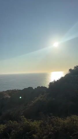 A video captures a wide view of a coastal landscape. The sun is visible in the upper right quadrant of the frame, casting a bright glare and a reflection on the calm ocean surface. The ocean stretches to the horizon, meeting a clear, pale blue sky. In the foreground and midground, a rugged, vegetated cliffside slopes down towards the sea. The vegetation appears dense and shrub-like, with some darker, bare patches of earth or rock visible. The lighting suggests it is either early morning or late afternoon. There are no people or discernable objects other than the natural landscape and the sun.