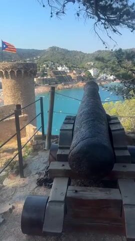 This video shows a view from a historical fortress overlooking a coastline. In the foreground, a large, weathered cannon mounted on a wooden carriage is prominently displayed. The cannon has a dark, rusted appearance and is secured with chains. Beyond the cannon, a stone wall and a round tower are visible. A flag, appearing to be the flag of Catalonia, flies from the top of the tower. The fortress walls extend along the hillside.

In the middle ground, a crowded beach with numerous sunbathers and umbrellas stretches along a bay. The water is a clear, turquoise blue and dotted with several boats. Buildings, some appearing to be hotels or apartments, are built on the hillsides surrounding the bay. The sky is clear and blue, indicating a sunny day. The overall impression is a scenic view of a coastal town with historical significance.