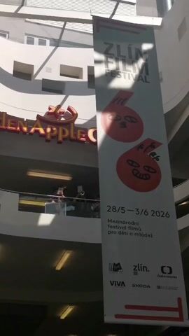 Here's a description of the video:

The video shows the interior of a multi-story building with a banner hanging that reads "Zlín Film Festival 28/5-3/6 2026 Mezinárodní festival filmů pro děti a mládež". The banner has two stylized red apple characters on it. To the left of the banner, there is a sign for "Golden Apple Cinema". On a balcony in the background, there are people, at least two of whom appear to be playing musical instruments. Light streams in from the ceiling.  The architecture is modern. There's a bit of glare from the sunlight.