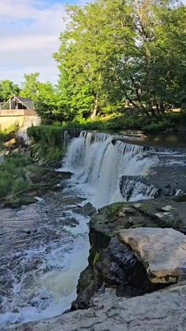The video shows a waterfall cascading down rocks into a body of water. Lush green trees and vegetation surround the waterfall, and a building is visible in the background. The water is white and frothy as it falls, and the surrounding rocks are wet and appear mossy in places. The sky is a pale blue with some clouds.