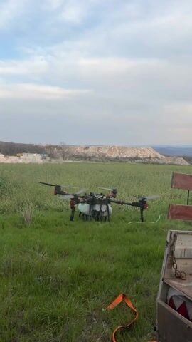 The video shows a drone taking off from a field. The drone is black and gray, with four sets of propellers. It is carrying a tank and nozzles, indicating that it is likely being used for spraying crops. The field is green and grassy, and in the background is a large pile of dirt or debris. There is a wooden fence on the right side of the frame, with an orange strap hanging over it. The sky is cloudy, suggesting an overcast day. The drone rises into the air, and the propellers spin rapidly.