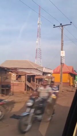 The video depicts a street scene in what appears to be a rural or developing area. A tall red and white telecommunications tower dominates the background. In the foreground are makeshift shops and stalls along the roadside. People are walking around, including one woman carrying a load on her head and a man riding a motorcycle. There are utility poles and power lines overhead. The overall scene suggests a bustling, but somewhat impoverished, environment.