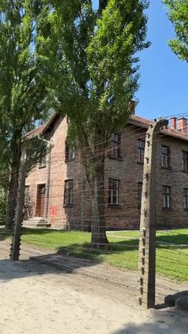 This media file shows a view of a brick building with multiple windows and a red-tiled roof, likely a historical site. The building is surrounded by a fence made of barbed wire and supported by concrete posts. A tall tree with lush green foliage stands in front of the building, casting shadows on the ground. The scene is set outdoors under a clear blue sky, suggesting a sunny day. The ground appears to be a dirt path or road, with wooden planks in front of the building and a grassy area.

The building itself is constructed of red brick, and some of the windows are barred, hinting at a past that may have involved confinement. There are visible house numbers above the entrance, with one reading "23". A red sign is also affixed to the wall near the entrance. The overall atmosphere is somber, evoked by the presence of the barbed wire fence and the stark architecture, which is often associated with former concentration camps or prisons. The time of day appears to be daytime, given the strong sunlight and shadows. The location is Oświęcim, Poland, which is famously known as the site of the Auschwitz concentration camp.
