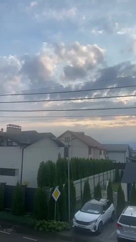 The video pans across a residential street with several houses and a row of tall, manicured evergreen trees lining a fence. Two cars, a white SUV and a silver SUV, are parked in a paved area. In the background, the sky is filled with clouds, with hints of sunset colors visible. Power lines are strung across the top of the frame.