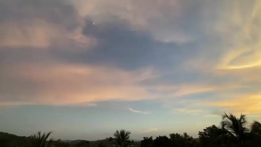 The video shows a time-lapse of a sunset over a tropical landscape. The sky is filled with wispy clouds that transition from soft pinks and oranges near the horizon to a pale blue at the top. A sliver of the moon is visible in the upper left quadrant of the sky. Below the sky, a silhouette of lush green foliage, including palm trees, forms the foreground. The landscape appears to be a rural area with a few distant hills. The lighting suggests the end of the day, with the sun setting beyond the visible frame. There are no people or animals visible in the footage.