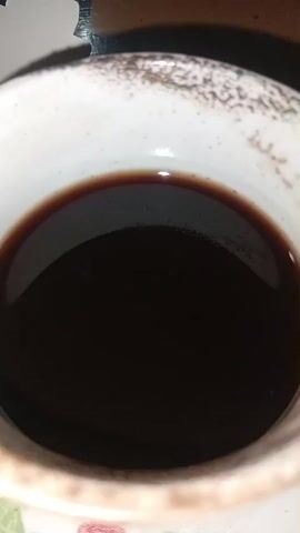 The video shows a close-up, top-down view of a dark liquid, likely coffee or tea, in a white ceramic cup with a speckled brown rim. The liquid has a slight sheen on its surface. The cup is positioned at an angle, revealing part of its textured exterior. The lighting suggests an indoor environment, with some shadows cast on the rim of the cup. There are no people or other objects visible in the frame. The video consists of still images that are rapidly presented, creating a slight visual flicker.