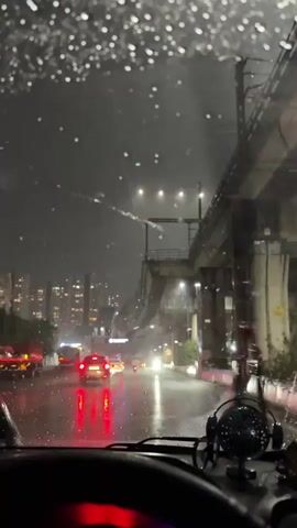 This is a first-person perspective video taken from inside a car during a heavy rainstorm at night. The windshield is covered in raindrops, and the windshield wipers are visible. The road ahead is wet and reflects the taillights of other vehicles, creating streaks of red light. Cars and trucks are driving on the road, their headlights and taillights illuminating the scene. In the background, tall buildings with lit windows are visible, suggesting an urban environment. An elevated structure, possibly a bridge or an overpass, runs parallel to the road on the right side of the frame. The overall atmosphere is dark and rainy, with the artificial lights from the city and vehicles piercing through the gloom.