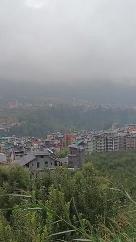 The video shows a slow pan across a mountainous, town with buildings scattered across the hillsides. The weather is overcast and misty, obscuring the tops of the distant mountains. The buildings are varied in style and color, with many appearing to be hotels or apartment complexes. There are also many trees and green foliage visible throughout the scene. The overall impression is of a peaceful, somewhat remote, mountain town.
