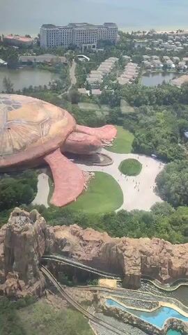 From a high vantage point, likely a building, the video shows a complex of structures. Dominating the view is a massive, stylized turtle structure, likely part of an amusement park or themed area. In the background is a hotel or resort complex near a beach. Below the turtle structure is a water park or attraction with a roller coaster-like structure and water features. People are visible on the ground near the turtle. The overall environment is lush and green. The video is a wide shot, offering a comprehensive view of the area.