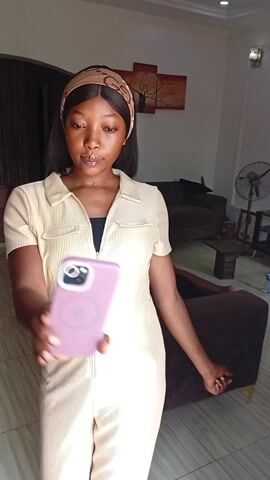 A young woman takes a selfie in a living room with loud music playing in the background. She stands near a doorway, wearing a beige dress, a brown patterned headband, and glossy lipstick. She holds her phone up to take the picture. In the background, there are brown couches, a dark wood end table, a floor fan, and a person reclining on the couch, partially obscured by a decorative pillow. A painting of a leafless tree hangs on the wall.