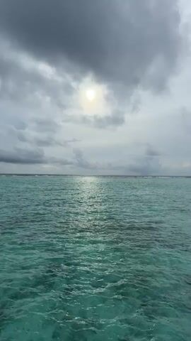 This media file captures a serene view of the ocean. The main subjects are the vast expanse of turquoise water and the sky above. The water appears clear and calm, with gentle ripples reflecting the light. In the distance, a thin line of white suggests a reef or a distant shore.

The sky is filled with dramatic, grey clouds, but a bright, almost luminous sun is visible peeking through a break in the clouds. This creates a striking contrast and casts a shimmering path of light across the water's surface. The overall mood is peaceful and contemplative, possibly evoking a sense of awe at the power and beauty of nature.

The time of day appears to be either sunrise or sunset, given the angle of the sun and the cloudy conditions that often accompany these times. The lighting suggests diffused daylight rather than harsh midday sun. The location cues, such as the clear turquoise water and the potential reef, strongly suggest a tropical or subtropical coastal setting, likely in a place known for its clear waters and coral formations. The Maldives is mentioned as a potential location, which aligns with these visual cues.

There is no visible text within the media file itself. The activity depicted is simply the static beauty of the ocean landscape. The gentle movement of the water and the play of light are the only signs of dynamism.