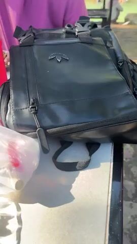 The video shows an outdoor scene with several people seated at tables. In the foreground, a person's hand is visible holding an object, possibly a phone. To the right, a black bag with a zipper is placed on a table. Next to it is a clear plastic bag containing food items. A pink water bottle filled with red liquid is also on the table. In the background, people are sitting at tables, some eating, under colorful umbrellas. The environment appears to be a recreational area, possibly a park or a resort, with trees and foliage visible. There are two women in the foreground, one wearing a pink hijab and the other a purple hijab, both seemingly engaged in eating. One woman is eating noodles with chopsticks, and the other is looking towards the camera. The overall atmosphere is casual and relaxed.