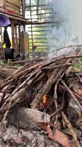 A close-up video shows a bonfire of dry twigs burning with an orange flame and releasing white smoke. Rocks surround the base of the fire. In the background, a simple wooden structure with a ladder and miscellaneous objects inside is partially visible. Beyond the structure, there is a green, tree-filled environment.