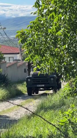 The video shows an old, green truck driving on a dirt road. The truck has a license plate visible that reads "HH-618-LL".  The road is lined with green grass and vegetation, and a tree partially obscures the truck's view. In the background, there's a house with a red-tiled roof and a view of mountains in the distance under a blue sky with white clouds. Electrical lines and a pole are visible to the left of the truck. The truck appears to be moving slowly, and the camera remains stationary.
