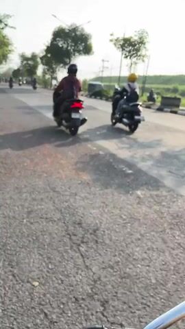 The video shows a road scene with several motorcycles and a car. The road surface appears worn and cracked. There are trees and utility poles along the side of the road. The sky is bright, suggesting it is daytime, possibly morning or late afternoon due to the quality of light. The camera appears to be mounted on a moving vehicle, likely a motorcycle given the perspective and slight shake. Two motorcyclists are visible in the foreground and mid-ground, both traveling in the same direction. One motorcycle has a rider wearing a helmet. Further down the road, another motorcycle is visible, and a dark-colored car is also present. The overall impression is of a typical scene on a paved road in an outdoor environment.