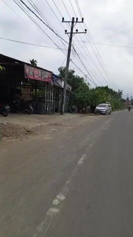 The video shows a gray van parked on the side of a two-lane road in an area with a mix of residential and commercial buildings, lots of wires overhead, and greenery. The camera is moving along the road, passing the parked van. There are other vehicles, including motorcycles, visible in the distance. The sky is overcast. The road appears to be in fair condition with some wear. There is a dirt shoulder to the left of the road, and vegetation is present along the roadside. A small wooden structure with an advertisement is visible to the left of the parked van.
