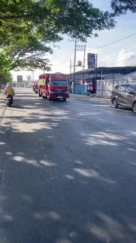 The video captures a street scene in what appears to be a town or city. The camera is moving forward, giving a first-person perspective. The road is paved and there is a significant amount of tree shade on the road surface, indicating a sunny day with trees lining the street.

In the foreground, the road is visible, with distinct tire tracks and shadows. As the camera moves, the scene unfolds.

On the right side of the road, a red truck with a cargo bed is prominently featured. It has decorative lights along the top and is driving away from the camera. The truck appears to be a commercial vehicle. Behind the truck, more buildings and shops are visible along the street. There are also utility poles and wires.

On the left side of the road, there are more trees, a wall, and parked cars. A person on a scooter is visible in the distance, also moving away from the camera. Further down the road, other vehicles, including motorcycles and cars, are present. The sky is blue with some clouds.

The overall impression is of a typical day on a street in a developed area, with traffic and commercial activity. The video is shot with a mobile device, likely a smartphone, given the framing and resolution. There is no audio present in the provided description.