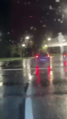 The video depicts a street scene at night during a heavy rain. The recording is taken from the inside of a vehicle with raindrops visible on the windshield. A blue car is stationary in the distance, its taillights reflecting vividly off the wet road surface. A motorcycle pulls up beside it. In the background, a white bridge-like structure is illuminated with lights. Streetlights line the road. A few pedestrians can be seen on the sidewalk. The sound of an engine can be heard in the background.