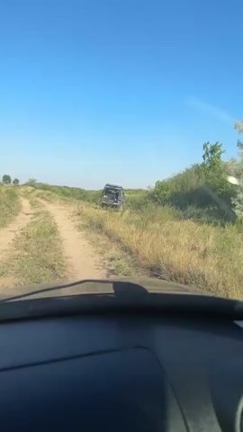 The video begins with a view from inside a vehicle, looking out at a dirt road winding through a grassy, rural landscape under a clear blue sky. A dark-colored SUV is visible in the distance, off to the right side of the road, partially obscured by tall grass. The camera pans slightly to the right, showing more of the overgrown vegetation and a distant treeline. The interior of the vehicle is then shown, focusing on the dashboard and then a man wearing sunglasses and a grey shirt, driving the vehicle. He glances to his left, away from the camera. The video continues to show the driver and the passing scenery of dry grass and trees.
