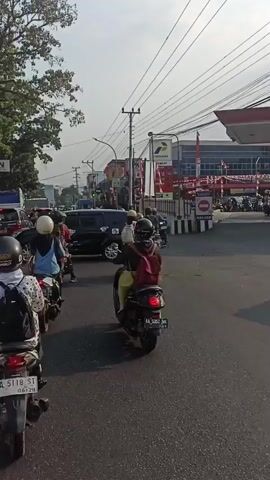 The video shows a street scene with several motorcycles and cars. The motorcyclists are wearing helmets and some have backpacks. The vehicles are on a paved road, and there are buildings and trees in the background. There are power lines and street lights visible. The traffic appears to be moving slowly, suggesting it might be congested. The scene appears to be in an urban or suburban area.