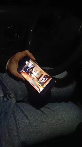 The video shows a person in a car holding a phone, which displays a scene from a game or animation of a man standing in an office, looking at another man seated at a desk. The scene is brightly lit on the phone screen. The car interior is dark and out of focus with a view of the steering wheel, dashboard, and red and green lights that are briefly visible on the right. The person is wearing blue jeans.