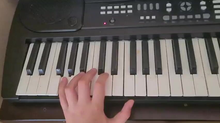 The media file shows a close-up shot of a person's left hand playing a black electronic keyboard. The keyboard has white and black keys. The person's fingers are positioned to play several keys. The activity is playing music on the keyboard. The scene appears to be indoors, with the lighting suggesting it could be daytime or a well-lit room. There are no clear indicators of the specific location, time of day, weather, or emotions. Some text is visible on the keyboard console, but it is too blurry to read. The overall setting is a person engaged in playing a musical instrument.