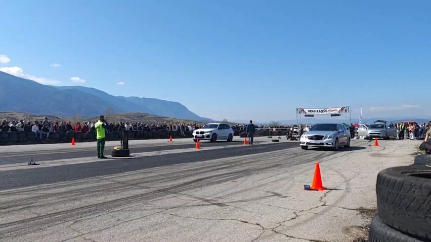 A silver Mercedes-Benz sedan and a white BMW X3 SUV are positioned at the start of a drag race track, side by side. A track official in a neon green vest stands between the cars, ready to signal the start. The track, marked by orange cones, leads away from the camera, with tire marks visible on the asphalt. A crowd of spectators lines the side of the track behind a barrier of tires. In the background, a mountain range stretches across the horizon under a blue sky dotted with some clouds. As the race starts, the Mercedes and BMW speed down the track. The BMW appears to accelerate faster. The video ends with the cars progressing towards the horizon.