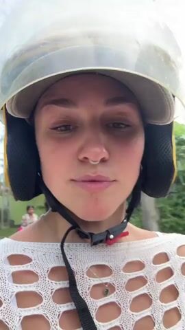 This media file features a close-up, point-of-view shot of a young woman riding on the back of a motorcycle in Kabalana, Sri Lanka. She is wearing a yellow motorcycle helmet with a clear visor, a white crocheted off-the-shoulder top revealing a white bikini top underneath, and light-colored pants. Her hair is pulled back, and she has a nose ring.

The background shows a blurry, outdoor scene with lush green foliage and what appears to be a building with a green wall and a tiled roof. The lighting suggests it is daytime, likely morning or afternoon, with no indication of specific weather conditions beyond being outdoors.

The woman's expression shifts from a neutral gaze in the first few frames to a broad smile as she looks to her left and then forward. Her eyes are closed briefly at the very beginning. The dynamic nature of the shot, with the slight shake and the motion blur of the surroundings, indicates the motorcycle is in motion. The presence of another person's dark clothing or hair is visible to the right of the frame, confirming she is a passenger. The overall impression is one of enjoyment and a carefree experience of traveling through a tropical environment. There is no visible text in the media.