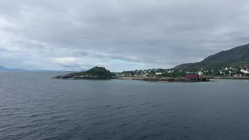 This video shows a panning shot of a coastal village in Norway. The water in the foreground is dark and choppy, suggesting a cloudy day. The land in the background features a small, rocky island with a lighthouse, followed by a cluster of houses and buildings lining the shore. Further inland, rolling green mountains rise, with some rocky outcrops visible. Several boats are docked in the harbor, and the overall impression is one of a remote, picturesque settlement. The sky is overcast with grey clouds.