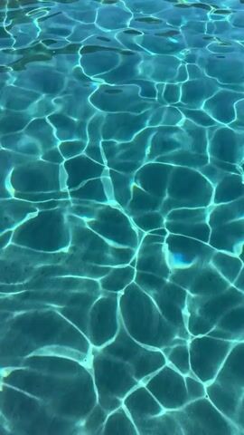 The video shows a close-up view of the surface of clear, turquoise water. Sunlight refracts through the water, creating shimmering patterns of light and shadow that move across the surface. The water appears calm, with gentle ripples. In the distance, a rocky shoreline with sparse vegetation is visible, along with a small white boat.