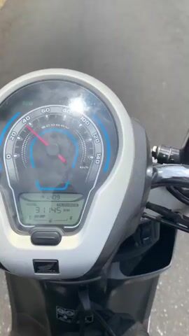 The video begins with a close-up of a scooter's dashboard. The speedometer shows the needle around 40 km/h, and the digital display shows the time as 12:09 and the total distance as 31145 km. The fuel gauge is full. The scene then shifts to a view from the scooter, showing a paved road winding through a lush green landscape. Hills covered in trees and vegetation rise on either side of the road, with a cloudy sky above. A few power lines are visible stretching across the frame. The scooter appears to be in motion, as evidenced by the slightly blurred background and the moving perspective.