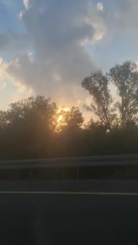 The video captures a sunrise over a landscape. The sun is a bright, glowing orb partially obscured by clouds and silhouetted trees. The sky is a mix of blue and white with large, fluffy clouds. In the foreground, there is a dark, horizontal line that appears to be a road or highway. The light from the sun creates a hazy, golden glow across the entire scene, giving it a soft and ethereal quality. The overall impression is one of a tranquil and beautiful dawn.