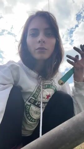A young woman with brown hair, wearing a white hoodie and black leggings, is crouching outdoors against a cloudy sky. She is holding a small gardening tool with a green and yellow handle in her right hand. Her hands appear dirty. In one shot, she holds the tool up and makes a peace sign with her left hand. She smiles at the camera.