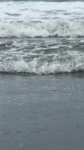 This is a close-up shot of ocean waves crashing onto a sandy beach. The water is a dark gray color, and the waves are frothy and white. The camera is positioned low to the ground, giving the viewer a perspective as if they are standing in the shallow water. The waves are breaking and receding, creating a dynamic and rhythmic movement. The sand is dark and wet from the water. The sky is not visible, and the focus is entirely on the interaction of the water and the shore. There are no people or other objects in the frame.