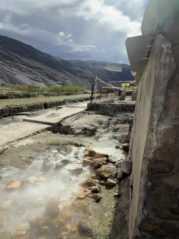 The video shows a geothermal spring with steam rising from the water. The spring is surrounded by rocks and concrete structures. In the background, there are mountains and buildings. The sky is cloudy. The camera pans from right to left, showing different angles of the spring and its surroundings.