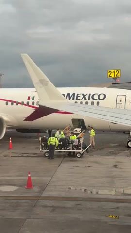 A commercial airplane with "AEROMEXICO" written on its side is parked at an airport gate. Several ground crew members wearing high-visibility vests are loading luggage onto the plane from a luggage cart. A green service vehicle is positioned to the left of the scene. Orange traffic cones are placed on the tarmac. The sky is overcast.