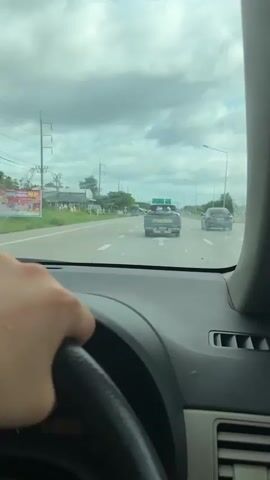 The video shows a driver's perspective from inside a car traveling on a multi-lane highway during daytime. A hand is visible on the steering wheel in the foreground, with the dashboard extending across the bottom of the frame. The windshield is slightly smudged.

Ahead on the road, a green Mazda pickup truck is visible, with two individuals covered in white cloth or blankets in its open bed. Another white pickup truck and other vehicles are further down the road. Overhead green road signs are prominent, displaying destinations in Thai and English: "พัทลุง" (Phatthalung), "ตรัง" (Trang), and "อ.ป่าพะยอม" (Pak Phayun/Pa Phayom). The sky is overcast with heavy clouds. Utility poles and power lines line the left side of the road, with trees and vegetation in the background. The text "Mazda" is clearly visible on the tailgate of the green pickup truck.